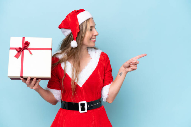 Young blonde woman dressed as Mama claus holding a present isolated on blue background pointing to the side to present a product - Photo, Image