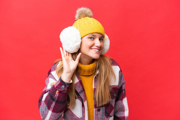 Молодая красивая женщина в зимних муфтах изолированы на красном фоне слушать что-то, положив руку на ухо - Фото, изображение