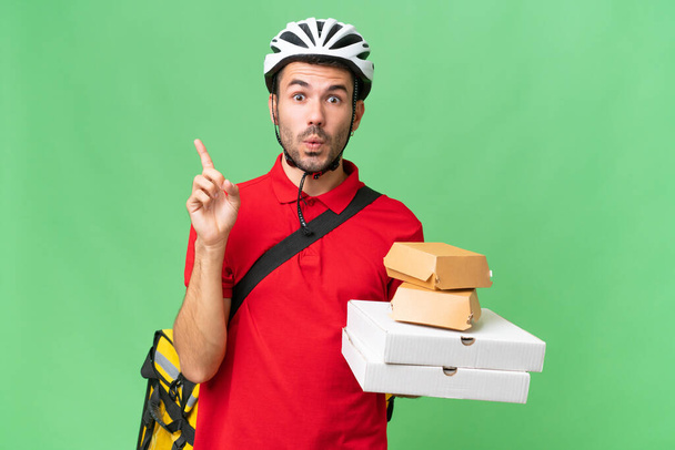 Young handsome caucasian man with thermal backpack and holding takeaway food over isolated background intending to realizes the solution while lifting a finger up - Photo, Image