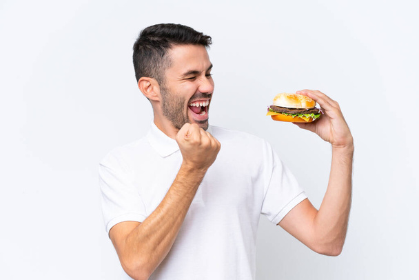 Fiatal jóképű kaukázusi férfi tartja a hamburgert az elszigetelt háttér ünneplő győzelem - Fotó, kép