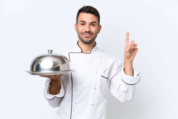 Jeune chef avec plateau isolé sur fond blanc montrant et levant un doigt en signe du meilleur - Photo, image