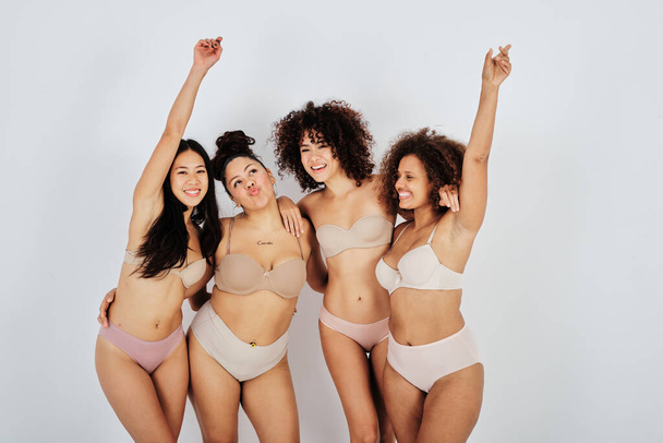 Delighted young slim and overweight multiethnic female models with dark hair in comfortable underwear smiling and cuddling standing in white studio with raised arms - Foto, Imagem