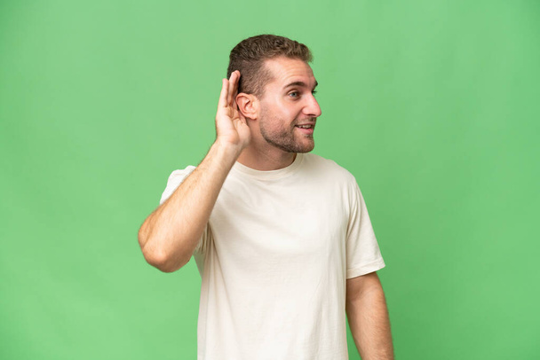 Fiatal jóképű fehér férfi elszigetelt zöld kroma háttér hallgat valamit azáltal, hogy kezét a fülre - Fotó, kép