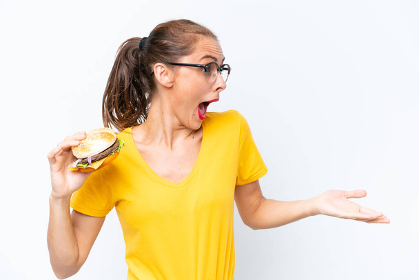 Young caucasian woman holding a burger isolated on white background with surprise expression while looking side - Foto, afbeelding