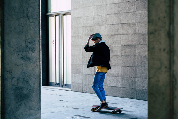 Side back view of one teenager boy using skateboard to move and transport. Green transportation and youthful lifestyle. Urban building style background. Modern guy using skate outdoor alone - Foto, Imagen
