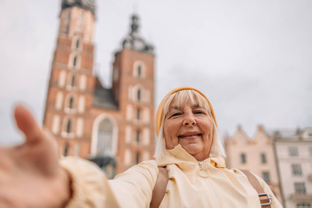 Portrait of 50s old woman with gorgeous smile with teeth and blonde hair against old city center view in Krakow. Takes selfie. - Photo, Image