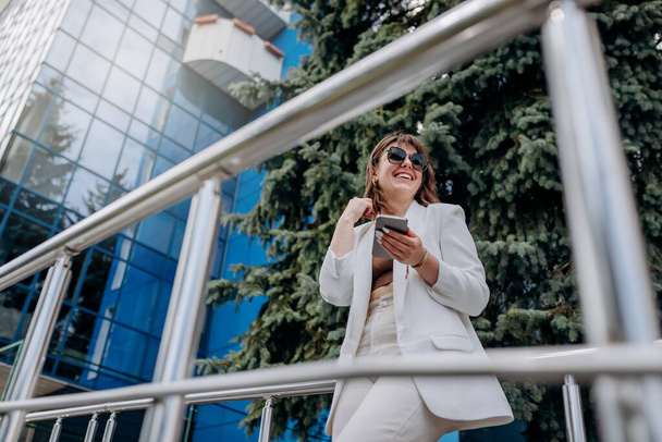 Lächelnde Geschäftsfrau in weißem Anzug und Sonnenbrille mit Handy in der Pause in der Nähe moderner Bürogebäude - Foto, Bild