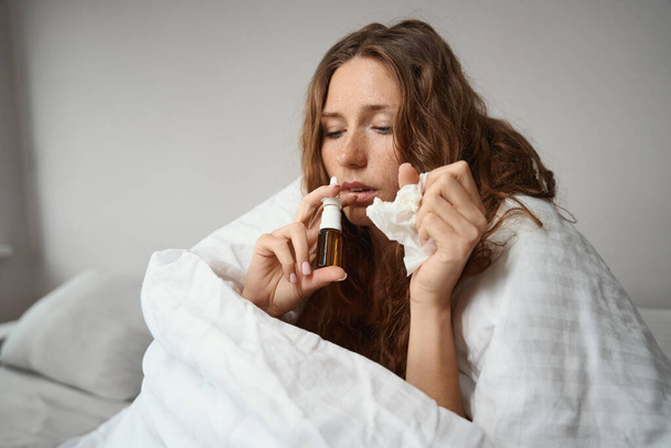 Sick lady wrapped in a blanket is feeling unwell due to a runny nose while in her hands is a treatment spray - Foto, Imagen