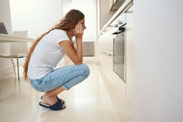 Thoughtful housewife sits in the kitchen near the oven in home clothes while food is being prepared in the oven - Foto, Bild
