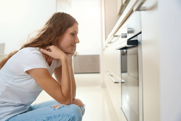 Thoughtful housewife crouched by electric oven while waiting for finished baking - Photo, Image