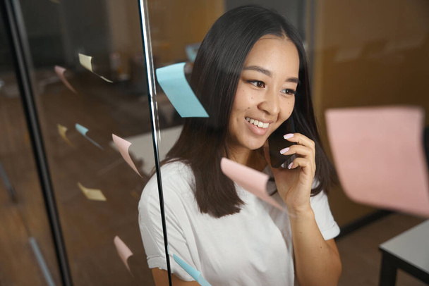 Close up portrait of smiling Asian female is having conversation on smartphone while standing near the glass door in the modern office - Photo, image