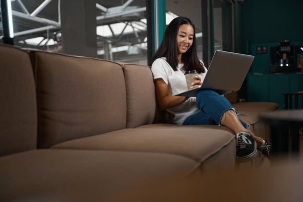 Full length side view portrait of happy cheerful Asian woman with coffee is sitting on the couch while reading something on computer in room indoors - Photo, image