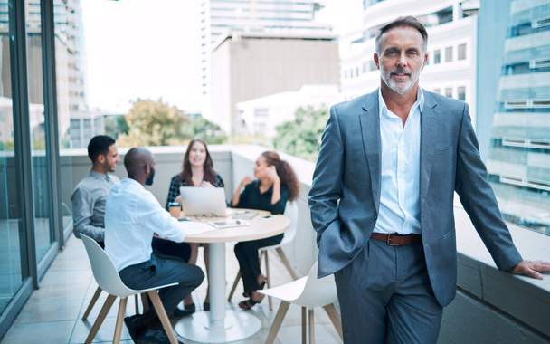 Чтобы стать лидером, нужна уверенность. Портрет уверенного взрослого бизнесмена, стоящего на балконе офиса со своими коллегами на заднем плане - Фото, изображение