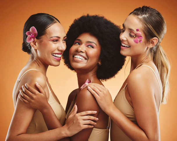 Beauty, flowers and women in makeup, diversity and models on orange studio background. Smile, skincare and happy, young and elegant female friends posing together with orchids after spa treatment - Foto, Imagem