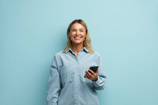 Happy Woman pitelee älypuhelinta eristyksissä. Valkoihoinen tyttö poseeraa matkapuhelimen kanssa, juttelee sosiaalisissa verkostoissa, käyttää mobiilisovellusta, tekstailee verkossa ystävien kanssa, jakaa uutisia, eristetty sinisellä  - Valokuva, kuva