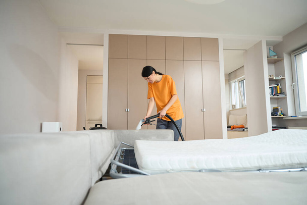 Woman in uniform standing in the bedroom near the sofa, holding vacuum cleaner and cleaning couch - Foto, afbeelding