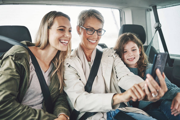 Phone, selfie and happy family in a car for a road trip driving or traveling to a holiday vacation adventure together. Grandma, mother and excited girl or child enjoy pictures for a fun weekend. - Photo, image