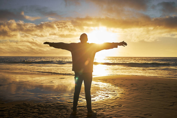 Поклоніння, схід сонця і силует людини на пляжі, що стоїть з піднятими руками. Віра, релігійна і духовна людина, дивлячись на сонце, океан і небо вранці, відчуває спокій, мир і надію в природі
. - Фото, зображення