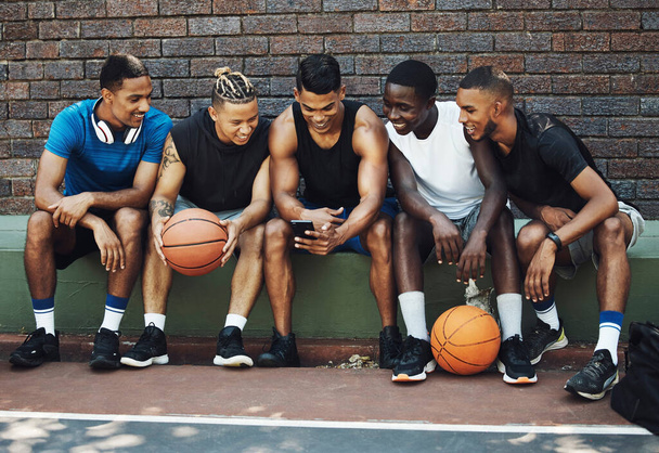 Basketballfreunde, ruhen Sie sich mit dem Handy aus und schauen Sie sich nach dem Training auf dem Sportplatz ein lustiges Video auf dem Smartphone an. Gesunde Männer, entspannt und müde auf der Bank nach dem Fitnessspiel. - Foto, Bild