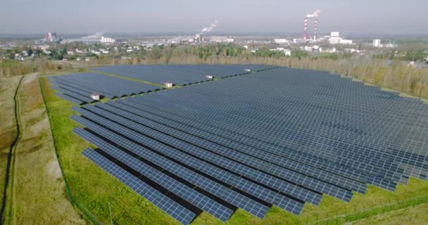 Los paneles solares se colocan en fila en los campos verdes cerca de la zona industrial con chimeneas. Fuente de energía ecológica renovable del sol. Vista aérea de filas de celdas fotovoltaicas, colectores solares. - Imágenes, Vídeo