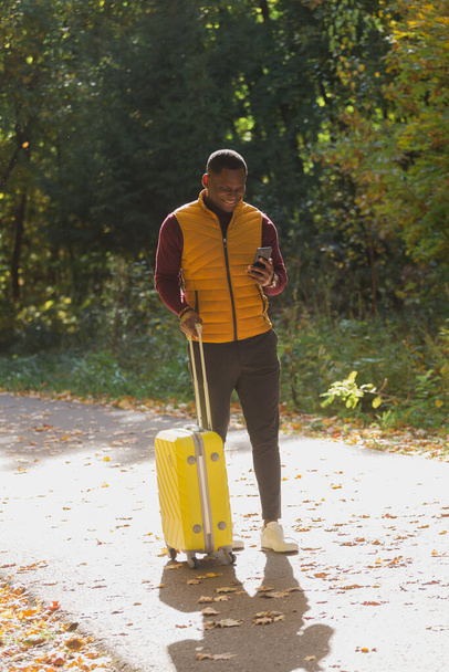 Joyeux jeune homme afro-américain en vêtements élégants avec valise se promène dans un parc d'automne par une chaude journée ensoleillée. Concept de voyage à la campagne - Photo, image