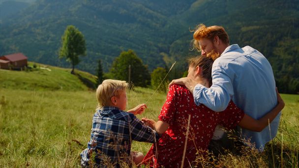 Loving parents hugging sitting with son on mountain slope sunny day. Relaxed family enjoying summer vacation on beautiful nature. Carefree couple relaxing on green hill with cute blond boy.  - Foto, Bild