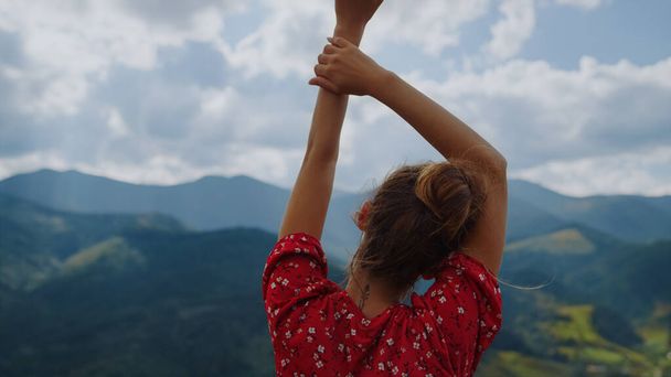Back view of unknown tranquil woman standing in front mountains scenery close up. Relaxed girl raising hands over head wearing red dress. Pretty lady enjoying summer holiday on cloudy nature. - Foto, Imagen