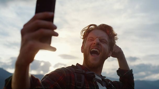 Closeup excited blogger take selfie on mobile phone outdoors. Hipster celebrate reach peak on mountains. Emotional traveling guy express happiness on digital smartphone. Technology leisure concept. - 写真・画像