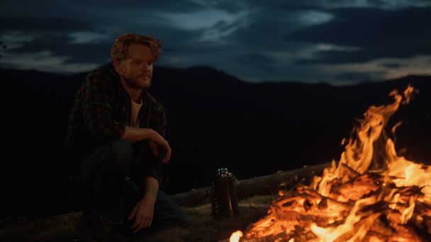 Dreamy hipster enjoy night fire burn on camp site. Closeup young traveler relax in mountains nature. Lonely millennial warm hands by campfire in dusk. Chill tourist rest on journey. Leisure concept. - Photo, image