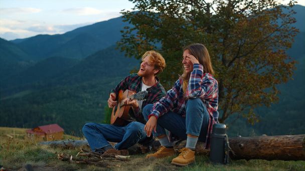 Cheerful couple enjoy nature at evening bonfire closeup. Young travelers play guitar in summer mountains. Smiling campers spend time together on vacation. Family holiday relax leisure travel concept. - Photo, Image