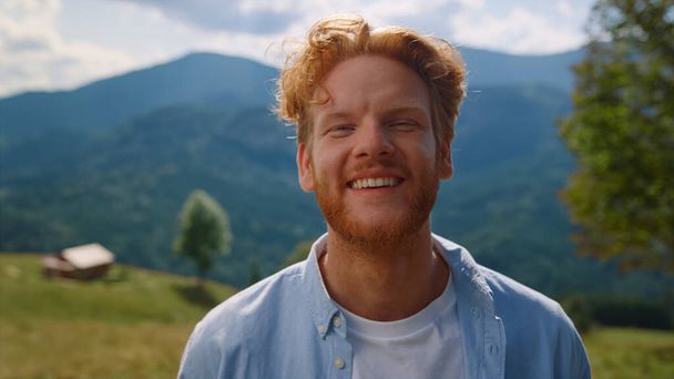 Portrait of happy man smiling on standing green meadow sunny day. Positive red hair guy relaxing in summer vacation outdoors. Cheerful person looking camera in front beautiful mountains close up. - Valokuva, kuva