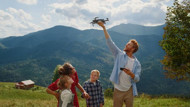 Happy family using drone on walk summer mountains. Red hair man launching quadcopter from hand outdoor. View of modern quadrocopter flying in cloudy sky operating by young guy. Technology concept. - 写真・画像