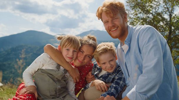 Happy parents posing with carefree children sitting green meadow sunny day closeup. Smiling family hugging looking camera in mountains. Cheerful young couple with cute kids enjoying summer holiday. - Foto, Imagem