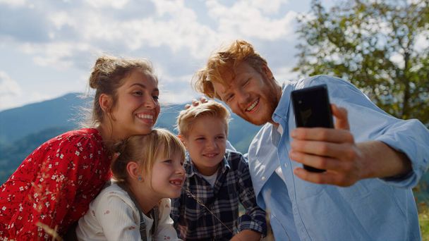 Ευτυχισμένη οικογένεια που έχει βιντεοκλήση στην ηλιόλουστη φύση. Χαμογελώντας νεαρούς γονείς με παιδιά που χρησιμοποιούν κινητό τηλέφωνο στα καλοκαιρινά βουνά. Χαρούμενο ζευγάρι με παιδιά που μιλούν στο κινητό τηλέφωνο κάθεται όμορφο λιβάδι. - Φωτογραφία, εικόνα