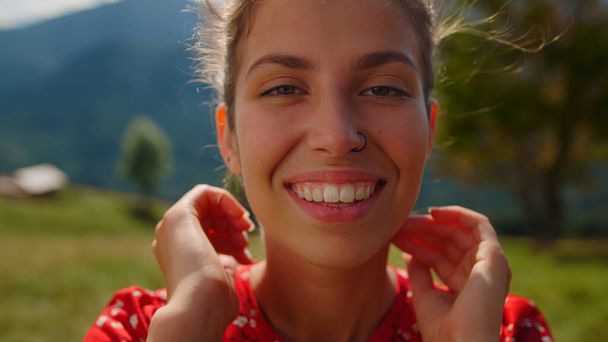 Πορτρέτο του χαρούμενου κοριτσιού χαμογελώντας κάμερα στο πράσινο λιβάδι. Χαλαρωμένη γυναίκα πρόσωπο μπροστά σε όμορφα βουνά τοπίο ηλιόλουστη μέρα. Κοντινό πλάνο θετικό μοντέλο θέτει στη φύση απολαμβάνοντας καλοκαιρινές διακοπές. - Φωτογραφία, εικόνα