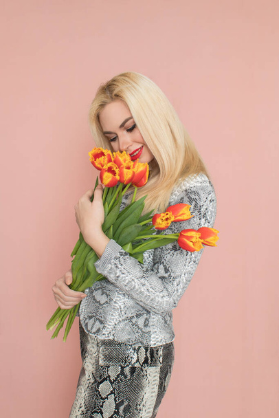Fashion photo of a beautiful elegant young woman in pretty snake suit, jacket blazer, top, skirt, massive chain around the neck posing on pink background. Studio shot. Bouquet orange tulips holding in hands. - Foto, Bild