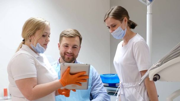 A handsome bearded young patient in the company of two female dentists looking at an Apple smartphone. Modern technologies of dental treatment - Zdjęcie, obraz