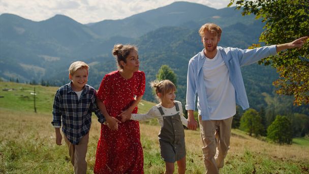 Happy walking family enjoying summer vacation on beautiful hill. Cheerful couple spending holiday with children in mountains. Joyful parents holding kids hands stepping green grass sunny nature. - Zdjęcie, obraz