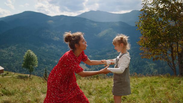 Happy mother enjoy funny dancing with daughter on green mountain hill. Cheerful woman teaching blonde girl dance element on nature. Pretty mom in red dress spending active summer holiday with child. - Photo, image