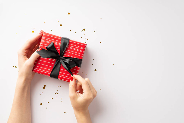 Black friday concept. First person top view photo of girl's hands untying ribbon bow on red giftbox over gold glowing confetti on isolated white background - Zdjęcie, obraz