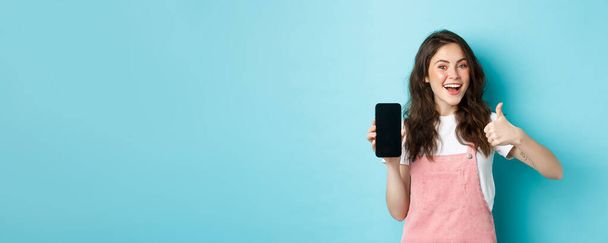 Так, це добре. Усміхнена мила дівчина показує великий палець і порожній екран смартфона, рекомендує програму або інтернет-магазин, стоячи на синьому фоні
. - Фото, зображення