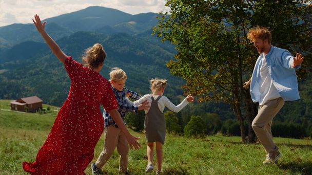 Cheerful family running like plane on mountain meadow. Playful people spreading hands wide imitating flight on green hill. Joyful parents playing with children outdoor games in summer vacation.  - 写真・画像