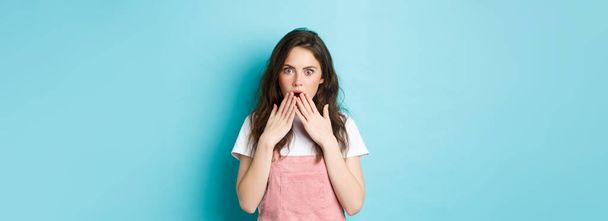 Portrait of shocked gossip girl gasping, covering opened mouth with hands and stare startled at camera, hear rumor, looking with disbelief, standing against blue background. - Foto, Bild