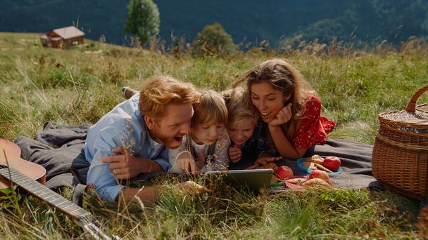 Positive family holding tablet computer on picnic lying green grass mountain hill. Smiling parents with children looking funny video on modern gadget outdoor. Happy people using tab at summer holiday. - Fotó, kép