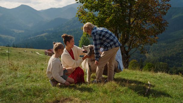 Cute family feeling happy with beloved dog on mountains slope. Smiling mother with blond son caressing adorable furry husky on summer walk sunny day. Happy people relax on nature with lovely pet. - Foto, Imagem