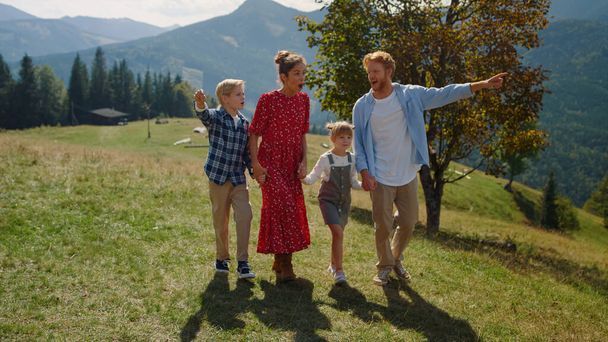 Smiling family enjoying nature walking on sunny mountain hill. Red hair man with cute blond son pointing fingers to mountain views outdoor. Happy parents talking with children going on green grass. - Photo, Image