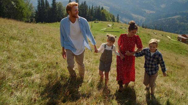 Friendly family walking on mountain slope sunny day. Happy parents holding children hands going to top green hill. Positive mother pointing finger on beautiful nature landscape. Vacation together. - Foto, Imagem