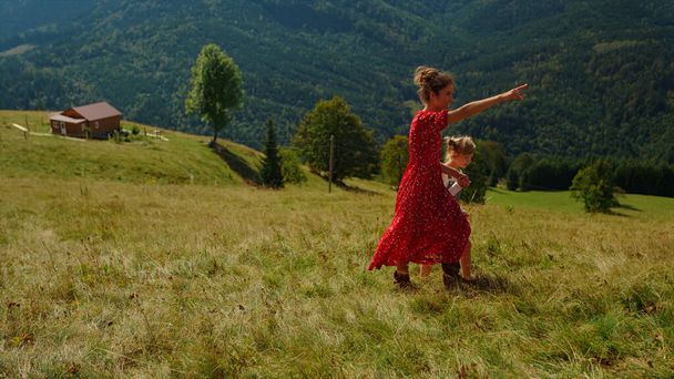 Happy mother walking with daughter on green grass mountain hill. Carefree woman pointing finger at nature views outdoors. Beautiful mom spending summer holiday with lovely child. Leisure together. - Fotografie, Obrázek