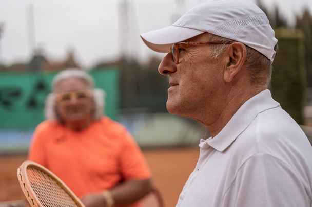 portrait of two senior tennis players dressed in sportswear relaxing at the end of the game on a clay tennis court - retired wellness concept - Foto, Bild