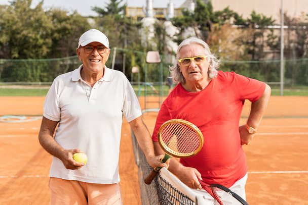 portrait of two senior tennis players dressed in sportswear shaking hands on a clay tennis court - retired wellness concept - Photo, Image